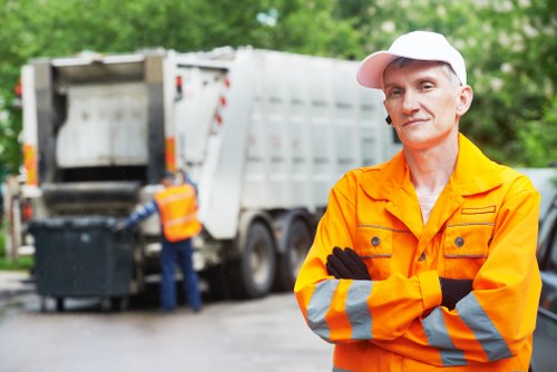 A professional waste management worker wearing an orange high-visibility jacket with reflective stripes and a white cap stands in the foreground with arms crossed, looking directly at the camera. Behind him, a large industrial waste disposal truck with an open rear compartment is actively involved in rubbish collection or emptying, with a worker partially visible at the back operating the vehicle. The scene takes place outdoors on a paved surface, surrounded by lush green trees, suggesting a suburban or urban environment in the vicinity of Alperton. The waste truck has a metallic finish with a slightly worn appearance, indicating typical use. The background is slightly blurred to emphasize the main subject, and natural daylight illuminates the scene, highlighting the details of the worker's attire and the vehicle. This setting illustrates a commercial waste removal operation in the local area, with Commercial Waste Removal Alperton providing rubbish collection services to businesses and local authorities.