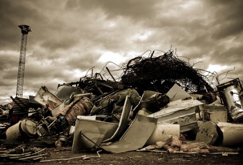 A large pile of discarded wreckage and debris is shown, consisting primarily of metal objects such as broken appliances, twisted steel frames, and various unidentifiable scrap materials. The debris appears weathered, with surfaces exhibiting rust and corrosion, and the colors range from dull greys and browns to patches of faded white and metallic finishes. The pile is situated outdoors on a flat, dirt-covered surface, with some items leaning or stacked unevenly, creating a chaotic arrangement. In the background, a tall metal structure resembling a communication or utility tower is visible against a cloudy, overcast sky, which adds a somber atmosphere to the scene. The environmental context suggests an industrial or junkyard setting, typical for waste disposal sites operated by companies such as Commercial Waste Removal Alperton, serving the local area. The overall image emphasizes the scale of metal scrap awaiting collection or processing, with minimal surrounding vegetation or structures, focusing on the heap of rubbish that a commercial waste removal service might handle in the Alperton area.