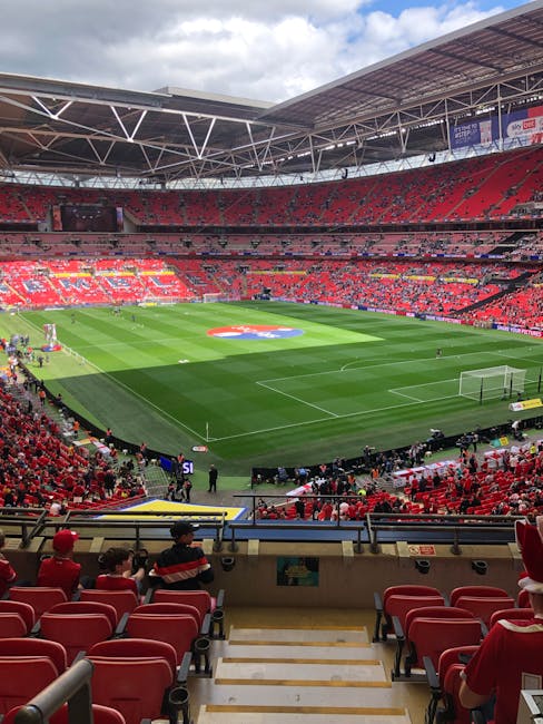 Inside a large football stadium with a partially covered, high arched roof, the view focuses on the empty, vibrant green grass pitch surrounded by red seating areas. The seating is mostly unoccupied,